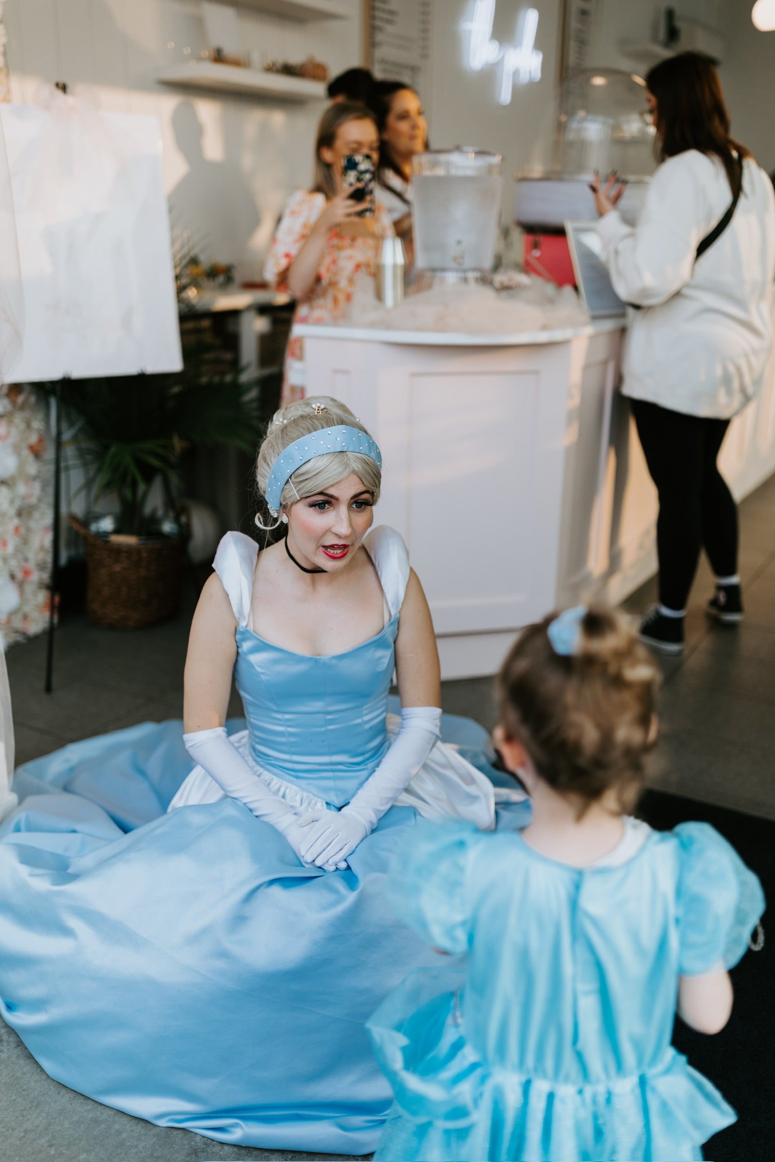Person in a blue dress sitting with a child in a similar dress in an indoor setting.