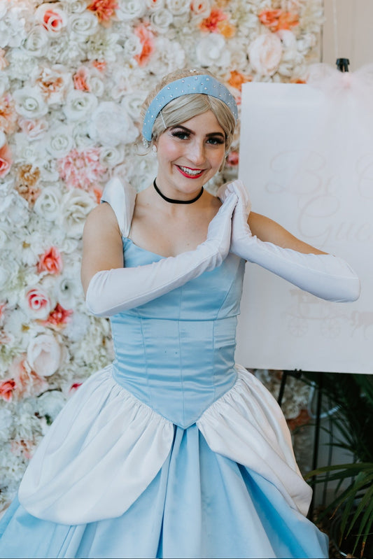 Person in a blue dress with white gloves posing in front of a floral wall.