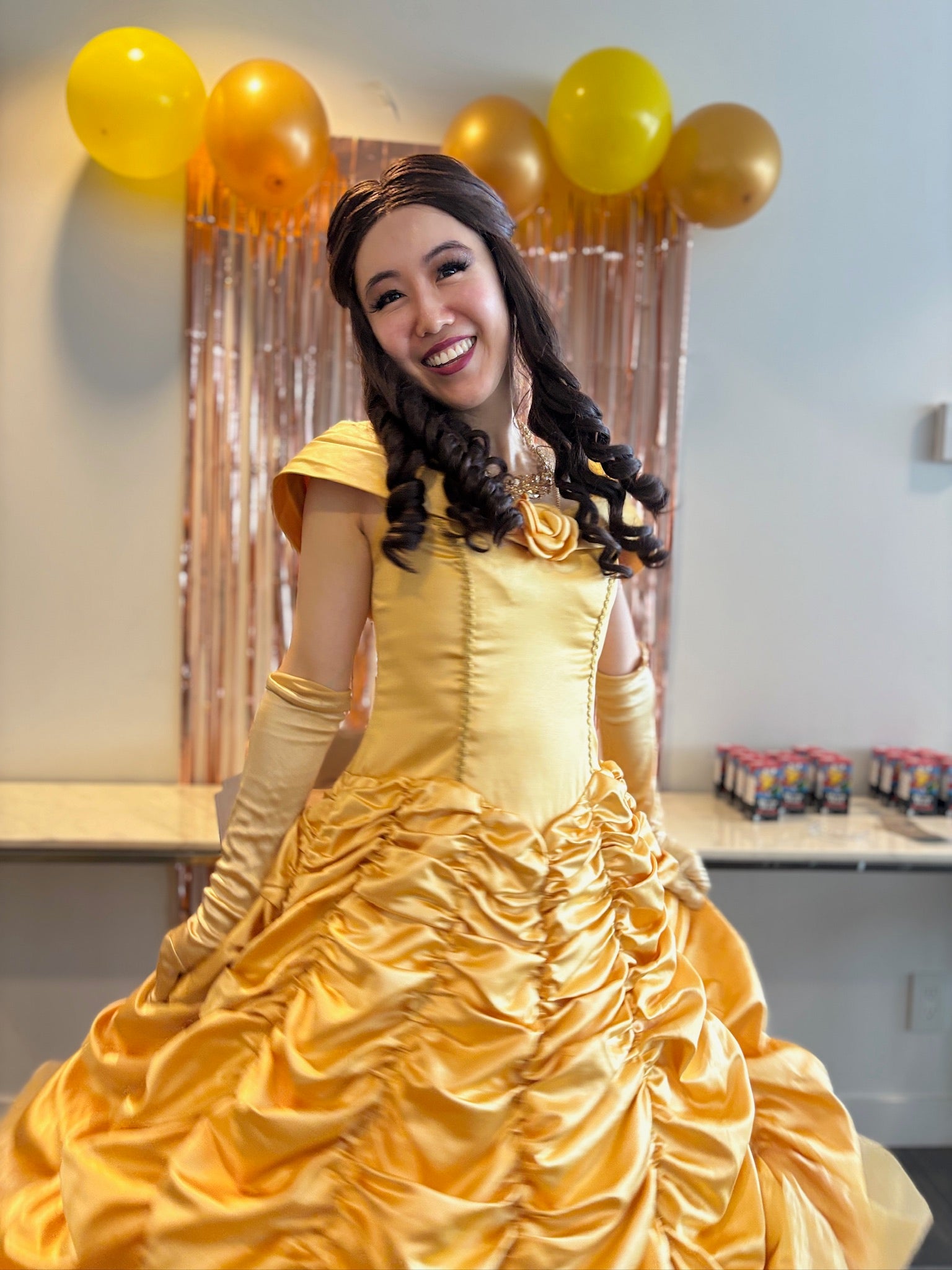 Person in a yellow dress with long gloves and braided hair, standing in front of balloons and a decorative curtain.